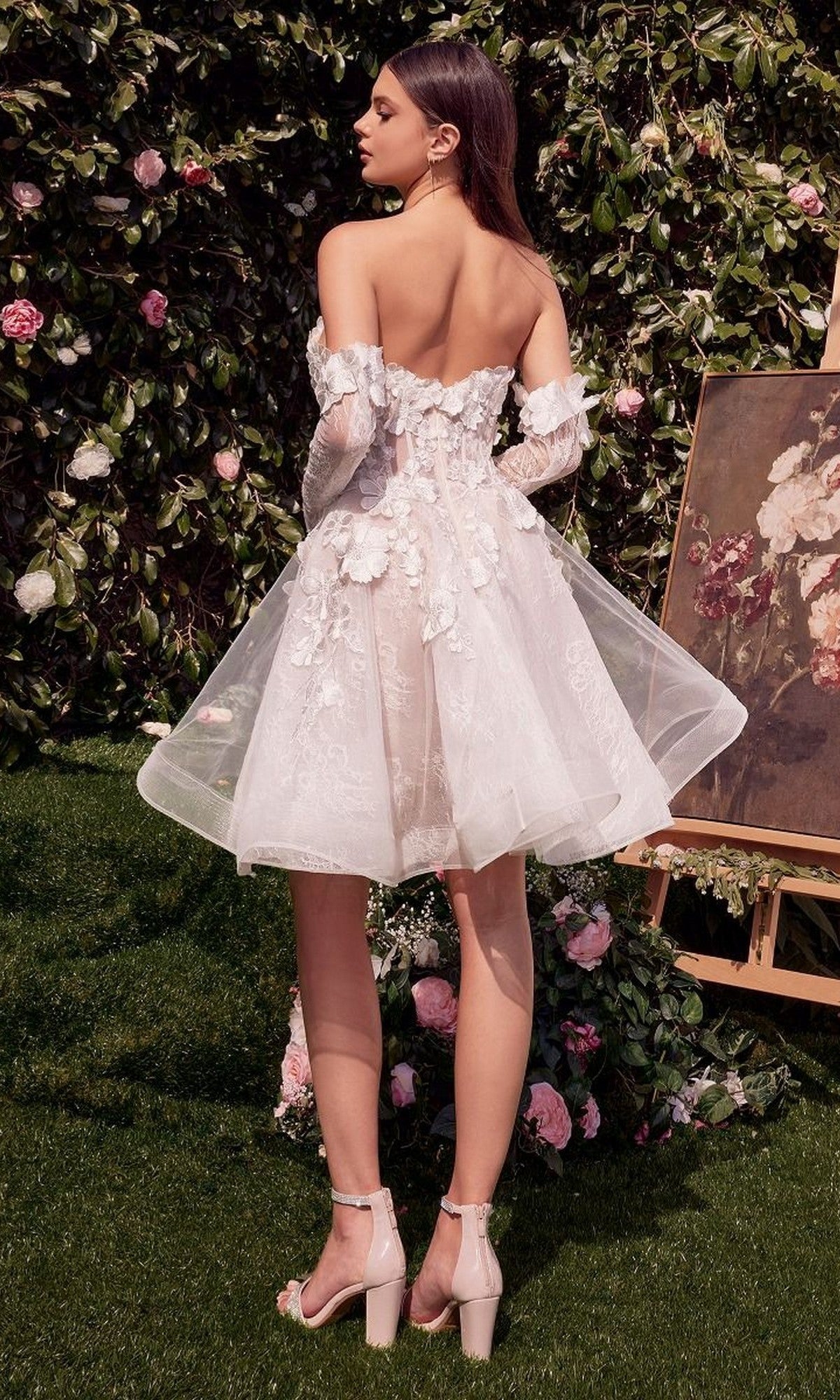 A woman stands outdoors in the Andrea and Leo WL020 Short White Party Dress with floral appliques and sheer sleeves, surrounded by greenery, flowers, and a nearby floral painting.